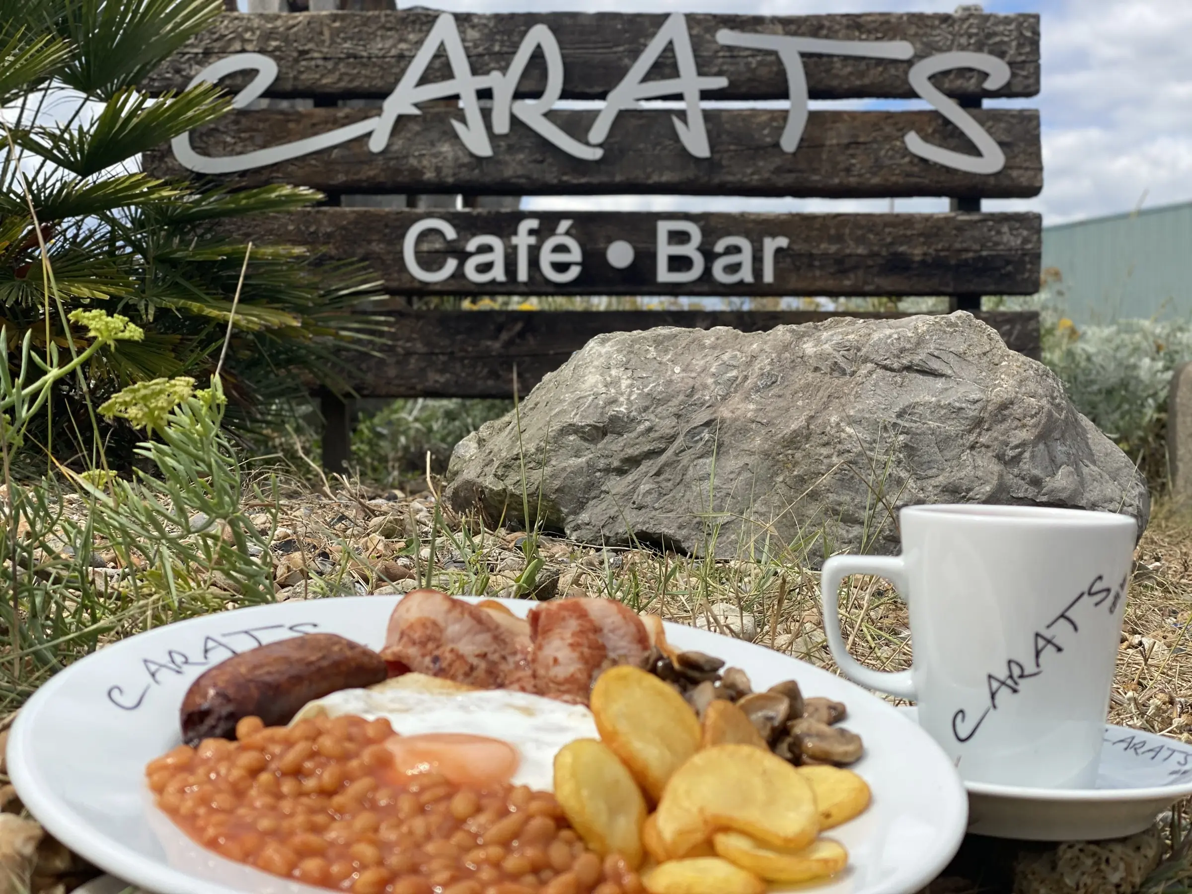 A full English breakfast and a mug of tea on Carats plates, in front of the wooden Carats Cafe Bar sign