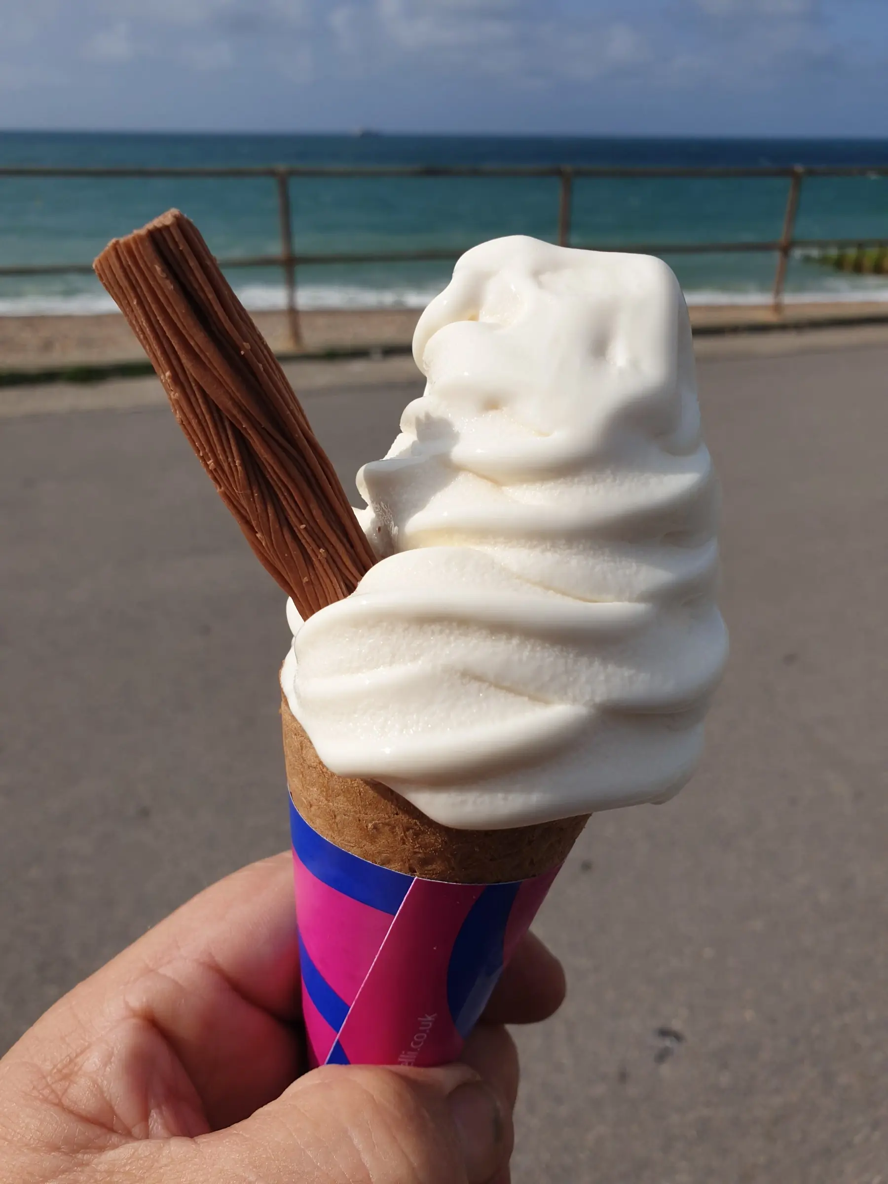 A soft-serve ice cream with a chocolate flake held up against the sea