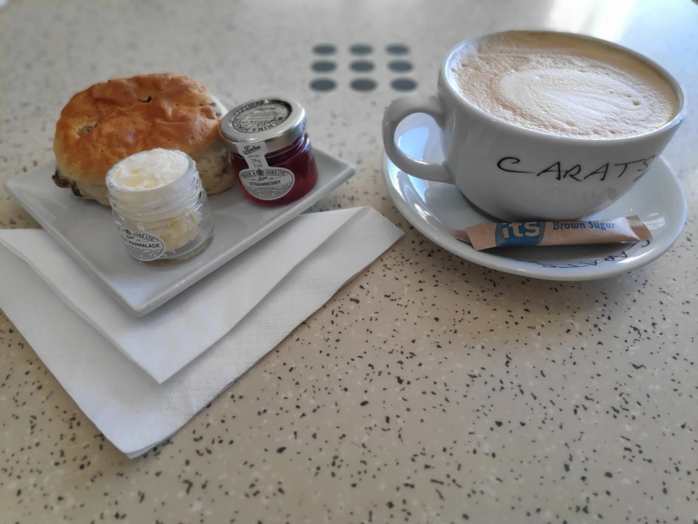A scone with jam and butter beside a cappuccino in a Carats cup
